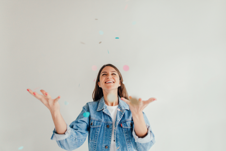 Image of a woman with brown hair in a denim jacket, white background. She has thrown confetti in the air & is smiling