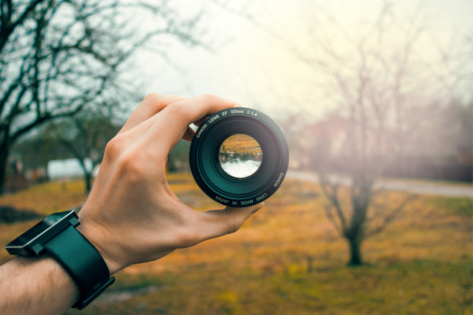 Image of a man's hand holding a camera lens out. The background is a park in late autumn/winter. A few trees with no leaves & the grass is a green/yellow