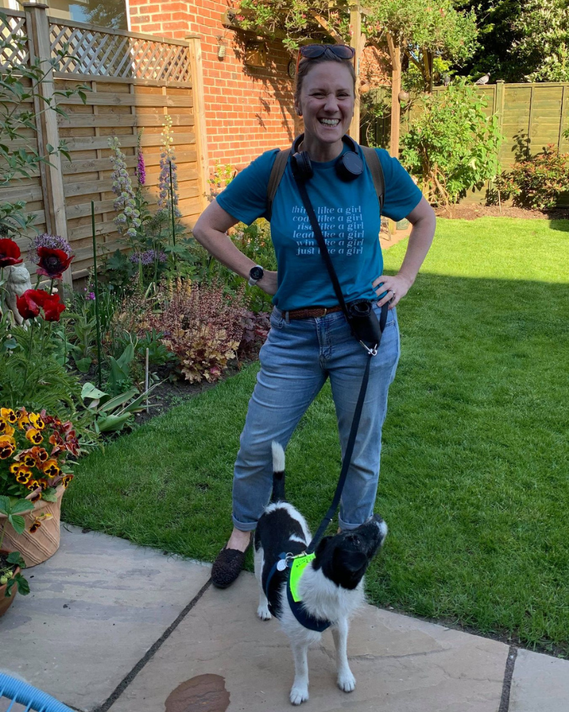 Image of hannah standing in a garden smiling/laughing, her dod Kiki is standing at her feet looking up at her.