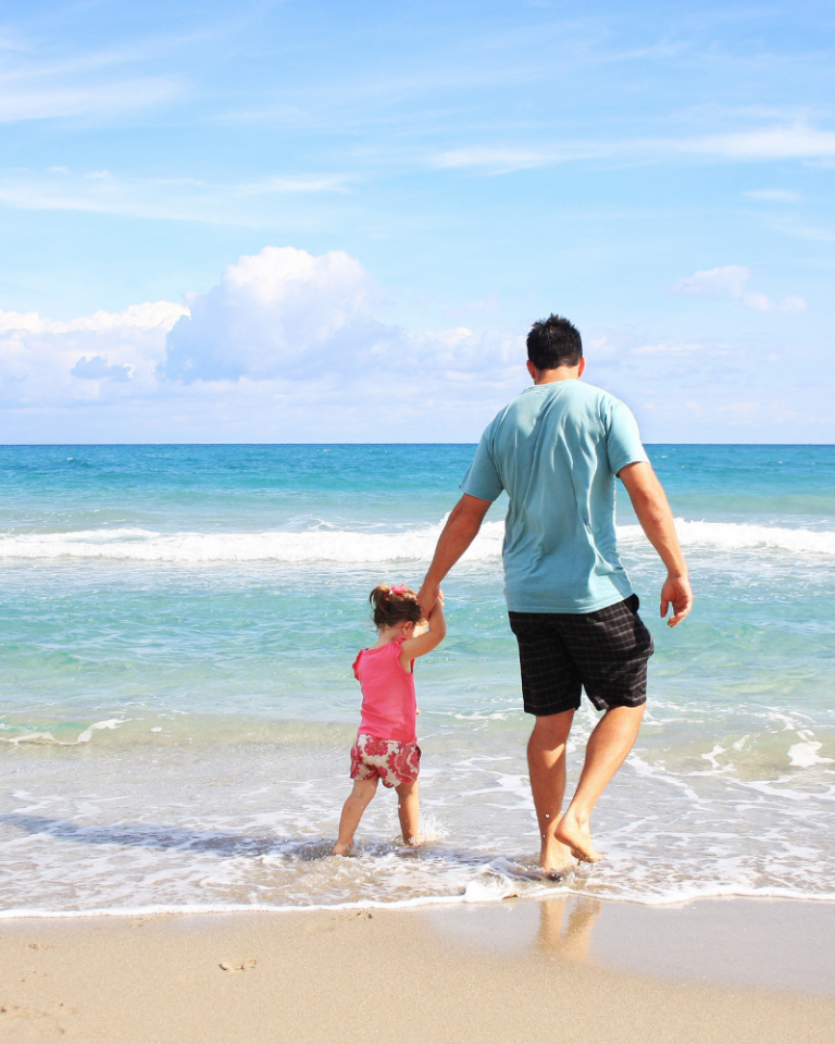 Image of a man holding a little girls hand paddling on what looks like a warm sunny beach