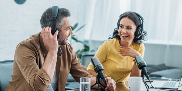 Image of a man and a women sat together at a desk recording a podcast, they are looking at each other & smiling