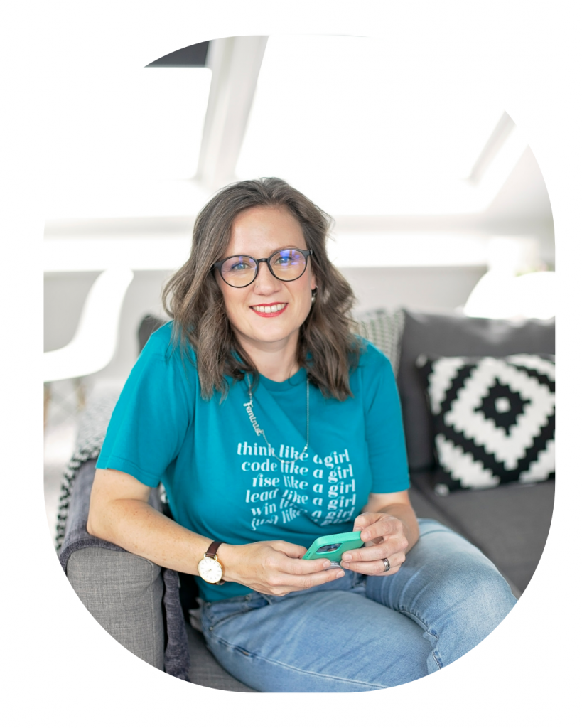 Image of Hannah, a white brunette wearing glasses. She is sitting on a grey sofa, holding her phone & looking towards the camera, smiling. She looks open and approachable.