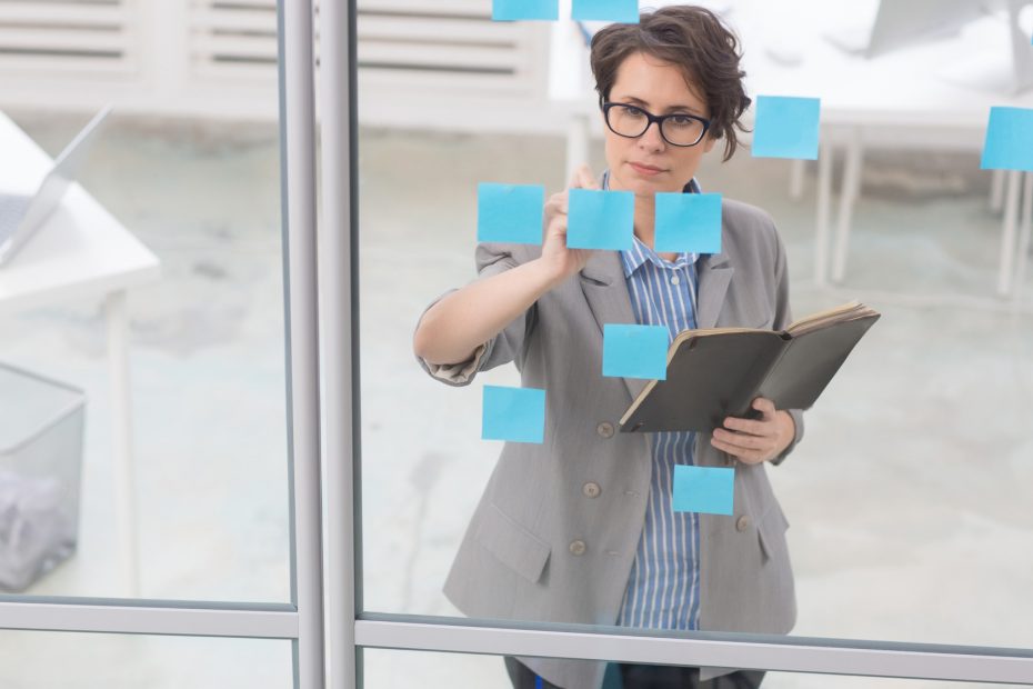 A white woman with short brown hair at a window with blue post-its on. She is writing on a post-it. Image conveys the person is creating an email workflow to make the most out of automated email tools