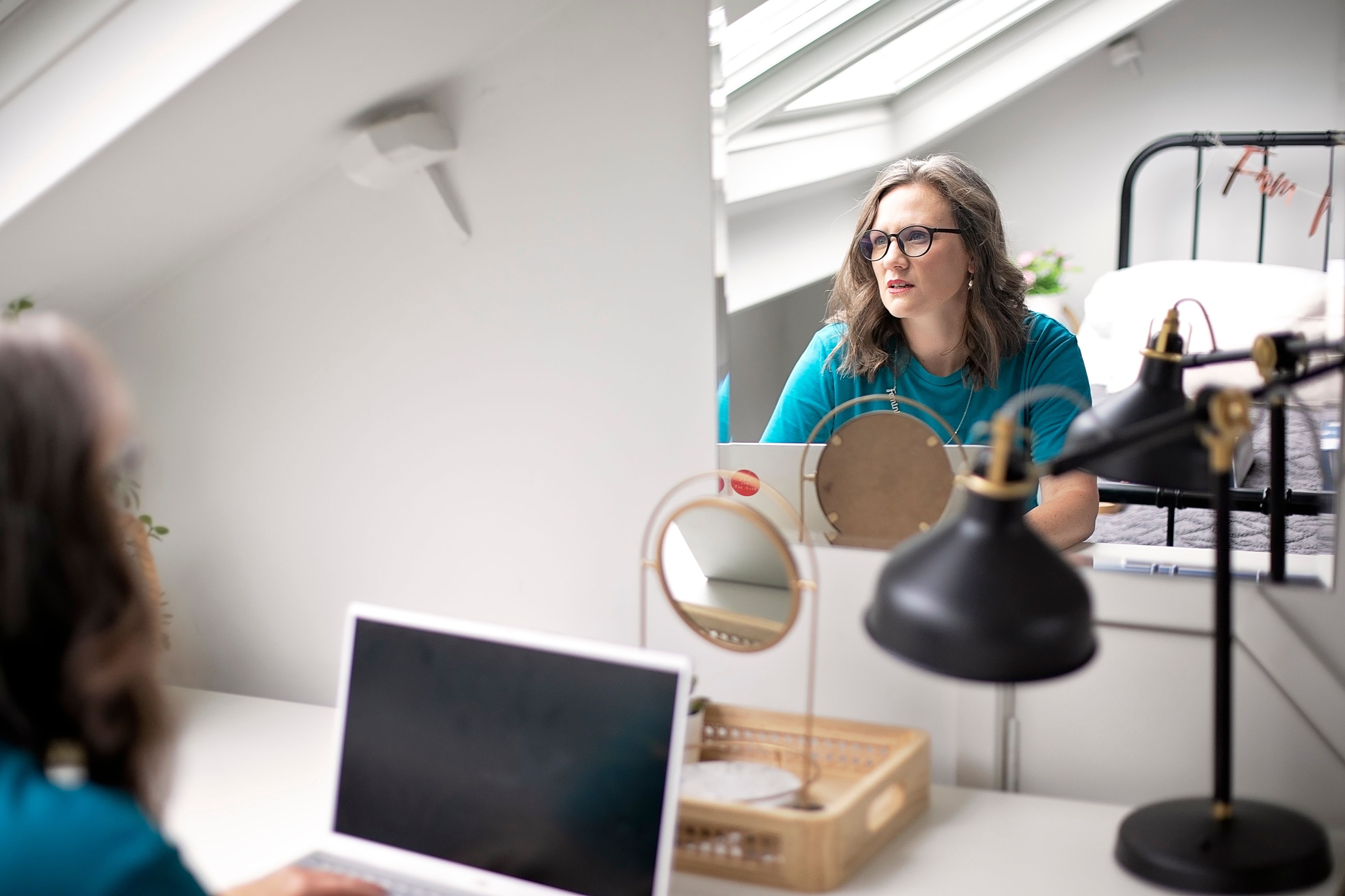 Image of a white woman with brown hair. Hannah is sitting at a desk staring into the distance, the photos taken from behind her but you can see her face in the reflection of the mirror. She looks confused as if trying to figure out MailerLite migration