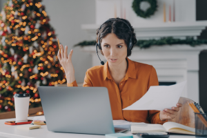 Image of a woman with a headset at a laptop with a Christmas tree in the background. Her hand is raised in the air. Image depicts the need to ease into the Holidays: Automate Your Business for a Relaxing Break.