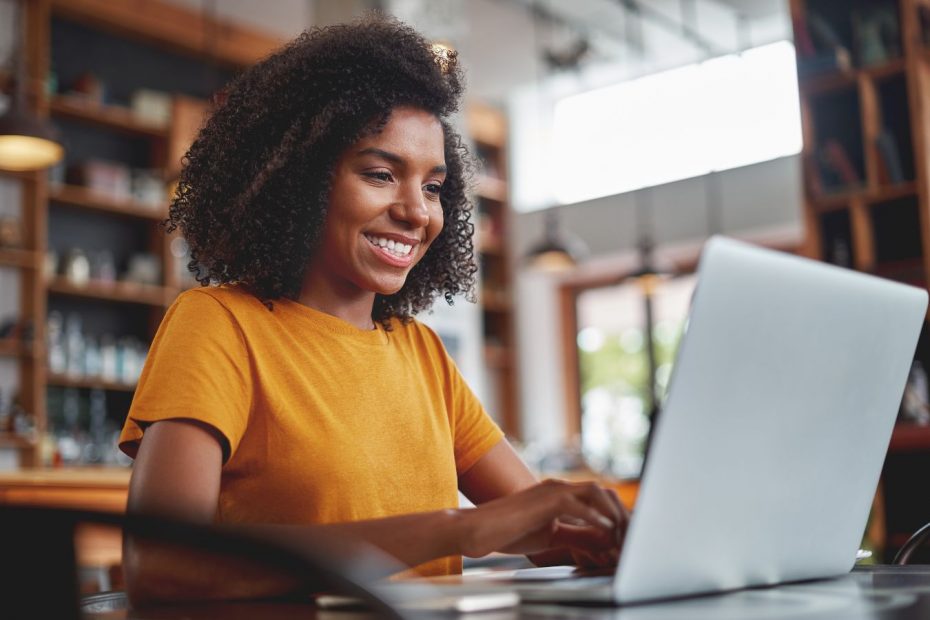 black woman with shoulder length brown curly hair sitting looking at a laptop and smiling. Image conveys that she is engaged with & enjoying the emails from the email funnel she is reading.