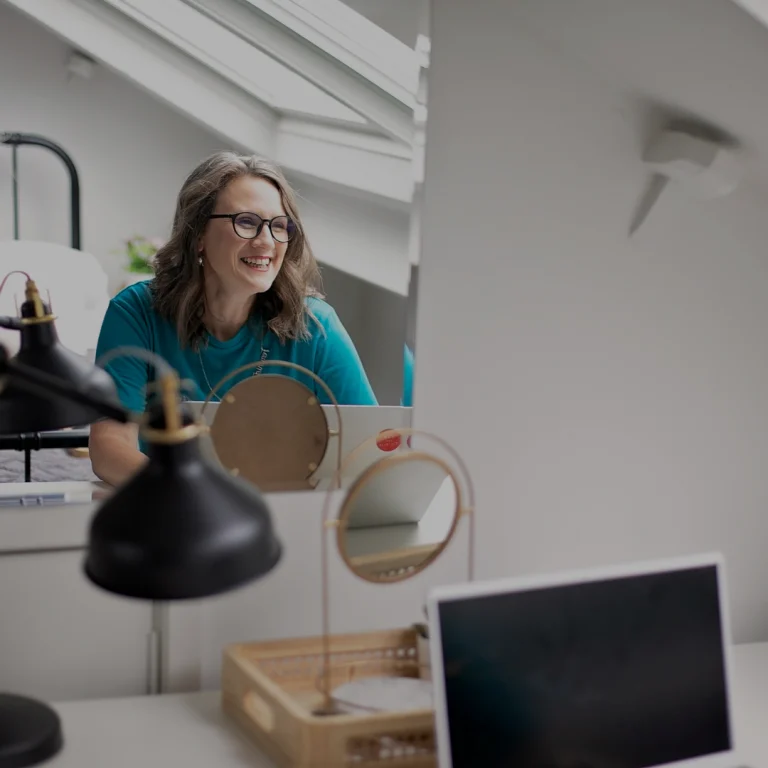 Image of Hannah Swierstra, white woman with brown hair, working at a desk with a mirror, the photo is taken form behind but you can see her reflection & she is smiling/laughing