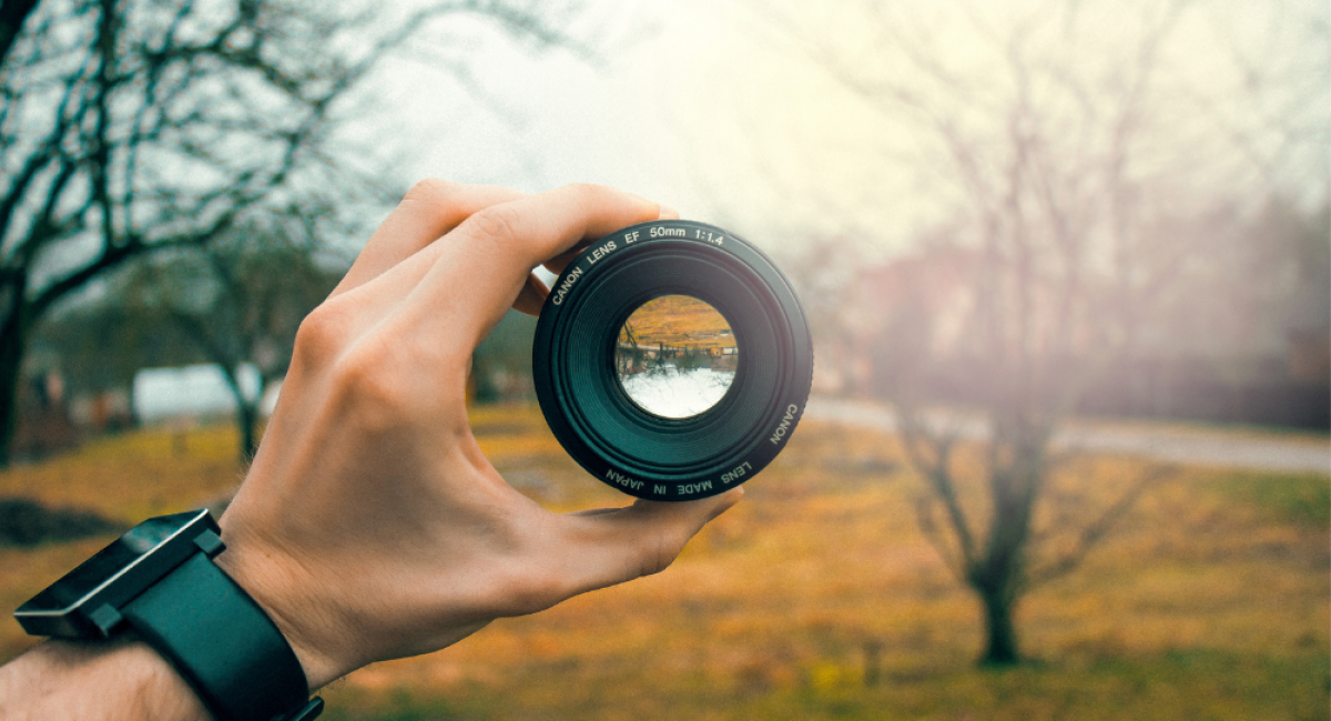 Image of a man's hand holding a camera lens out. The background is a park in late autumn/winter. A few trees with no leaves & the grass is a green/yellow